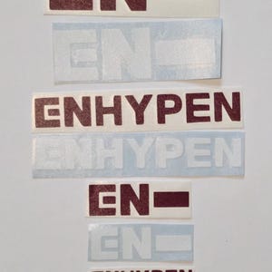 May include: A collection of stickers featuring the text "EN" and "ENHYPEN" in a burgundy color. The stickers are rectangular and have a white background. Some stickers have a burgundy rectangle next to the "EN".