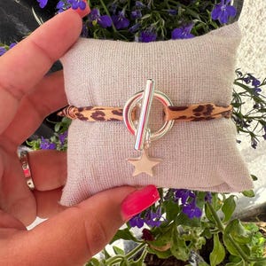 Può includere: Un braccialetto d'argento con un ciondolo a forma di stella e una fascia in tessuto con stampa leopardata. Il braccialetto è su un piccolo cuscino marrone chiaro.