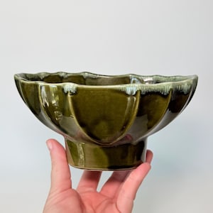 May include: A green ceramic bowl with a scalloped rim and a pedestal base. The bowl has a glossy finish and a dark green color with lighter green and white accents near the rim. The bowl is held by a hand.
