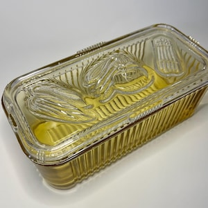 May include: A rectangular, amber-colored glass casserole dish with a clear lid. The lid features embossed corn and asparagus designs. The dish has a ribbed exterior and is set against a white background.