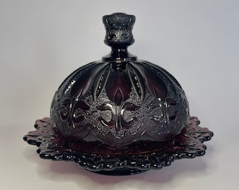 May include: A dark red glass butter dish with a domed lid and a decorative base. The lid features embossed floral patterns and a small handle. The base has a scalloped edge. The dish is likely vintage.