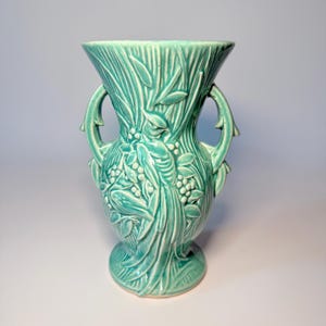 May include: A teal ceramic vase with a detailed relief design. The vase features a flared top, two curved handles, and a pedestal base. The relief depicts leaves, berries, and a bird, creating a nature-inspired aesthetic. The vase is approximately 9 inches tall.