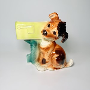 May include: A ceramic dog figurine with a mailbox. The dog is brown, white, and black, with a glossy finish. The mailbox is yellow and teal. The figurine is a decorative item.