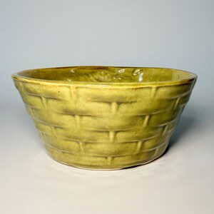 May include: A ceramic bowl with a glossy, chartreuse-green glaze. The exterior surface features a moulded, raised texture that mimics the appearance of a woven basket pattern, with horizontal and vertical lines creating a grid-like design.