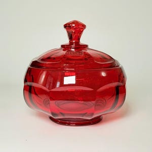 May include: A ruby red glass jar with a lid. The jar has a round, faceted body and a matching lid with a diamond-shaped handle. The jar sits on a small, round base. The glass is transparent, allowing light to pass through.