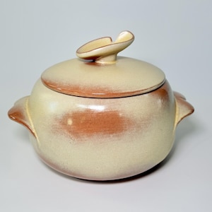 May include: A ceramic lidded serving dish with a cream and reddish-brown glaze. The dish has two handles and a decorative finial on the lid. The overall shape is round, and the design is simple and elegant.