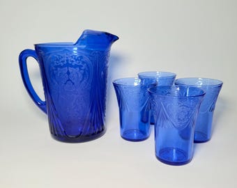May include: A cobalt blue glass pitcher and four matching tumblers. The pitcher has a handle and a spout. All pieces feature an embossed floral design. The glassware is suitable for serving water, juice, or other beverages.