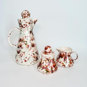 May include: A ceramic set consisting of a tall pitcher with a lid, a sugar bowl with a lid, and a creamer. Each piece features a white base with a multi-colored speckled pattern in shades of red, orange, green, and brown across the surface.