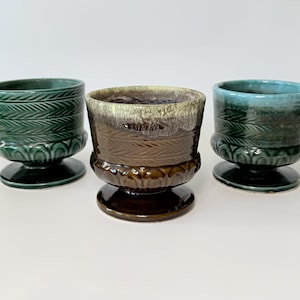 May include: Three ceramic pedestal planters in shades of green and brown. The planters have a textured, leaf-like pattern on the sides and sit on a round base. The center planter has a two-tone brown and cream glaze.