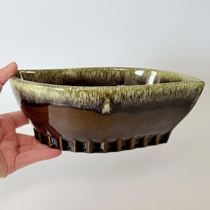May include: A brown and green ceramic bowl with a unique shape. The bowl has a glossy finish with a dark brown base and a lighter green rim. The bowl is held by a hand.