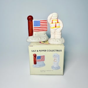May include: A pair of ceramic salt and pepper shakers. One is a red and white American flag, and the other is an astronaut. The shakers are on a box that reads "SALT & PEPPER COLLECTIBLES". The box also shows the images of the shakers.