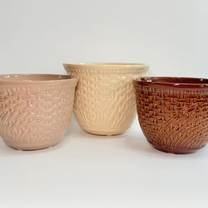 May include: Three ceramic planters with a textured basket-weave pattern on the exterior. The planters are arranged in a row, featuring a light mauve, a cream, and a dark brown glossy finish, each with a wide circular opening and a tapered base.