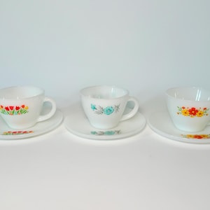 May include: Three white teacups and saucers with floral designs. Each cup and saucer features a different colorful floral pattern: red tulips, blue flowers, and yellow and orange daisies. The cups have a classic handle design.