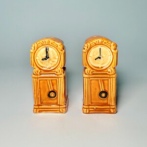 May include: Two ceramic salt and pepper shakers shaped like miniature grandfather clocks. The clocks are a warm brown color with cream-colored clock faces and black clock hands. Each clock has a circular pendulum.