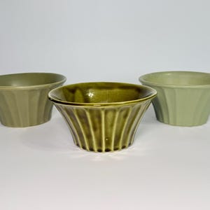 May include: Three ceramic bowls in shades of green are arranged on a white surface. The bowls have a fluted design and a flared shape. The center bowl is a darker olive green, while the other two are lighter shades.