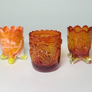 May include: Three antique glass toothpick holders in shades of orange and amber. Each holder has a unique design, including floral and patterned details. They are elevated on small, decorative feet, showcasing a vintage aesthetic.