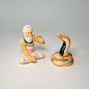 May include: A pair of ceramic figurines depicting a turbaned man and a cobra. The man is in a seated position, with a white turban, beard, and orange skin. The cobra is coiled, with a raised head and a black and brown pattern.