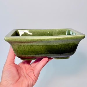May include: A rectangular ceramic bonsai pot with a glossy, mottled olive green glaze. The vessel features a wide, flared rim, slightly tapered sides, and four small, integrated feet at the base, showcasing a smooth, reflective finish.