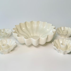 May include: A cream-colored ceramic serving bowl with a scalloped edge, accompanied by four matching candle holders. The bowl has a fluted design, and the candle holders have a similar scalloped shape. The set is displayed on a white surface.