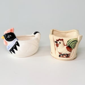 May include: Two ceramic hen and rooster-themed containers. The hen-shaped container is white with black, blue, and pink accents. The rooster-shaped container is beige with a rooster illustration in red, green, and brown.