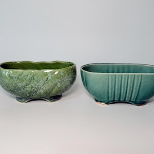 May include: Two oval ceramic planters. The left planter features a mottled moss green glaze with a wavy rim and a footed base. The right planter has a solid teal blue-green glaze, vertical ribbed detailing on the sides, and a footed base.