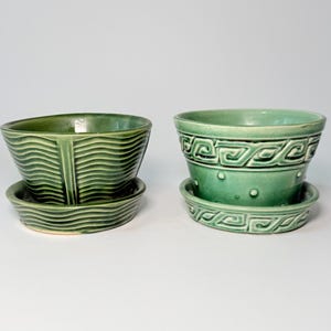May include: Two green ceramic planters with matching saucers. The left planter features a wavy, horizontal line texture. The right planter displays a geometric Greek key pattern around the rim and a series of small raised dots along the base.