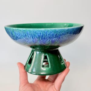 May include: A ceramic pedestal bowl with a glossy finish. The bowl's interior transitions from a deep teal to a vibrant blue, while the base is a solid green. The base features cut-out geometric shapes.