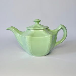 May include: A light green ceramic teapot with a lid. The teapot has a curved handle and spout. The body of the teapot has a textured design. The teapot is a pastel green color.