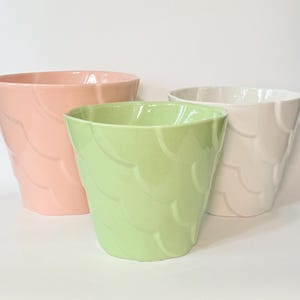 May include: Three ceramic planters in pastel colors: one light pink, one light green, and one white. Each planter features a repeating embossed scalloped or fish scale pattern on the exterior surface, with a wide, open top and a tapered base.