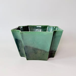 May include: A green ceramic bowl with a geometric, faceted design. The bowl has a glossy finish and a gradient of green hues, transitioning from a lighter shade at the top to a darker shade at the bottom. The bowl is empty and sits on a white surface.