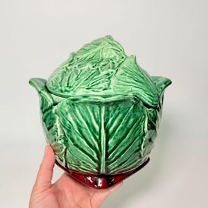 May include: A green ceramic cabbage-shaped serving dish with a lid. The dish has a glossy finish and is detailed with leaf textures. The base is a deep red color. A hand is holding the dish.