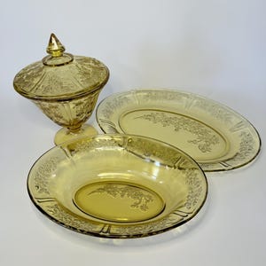 May include: A set of vintage amber-colored glassware, including a covered candy dish, an oval serving platter, and a round bowl. Each piece features a floral design. The glassware is likely made of pressed glass.