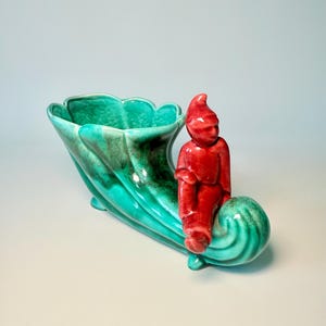 May include: A ceramic planter in the shape of a horn, with a teal and green glaze. A small red gnome figure sits on the side. The planter has a scalloped rim and a swirling design.