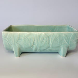 Peut inclure: Un pot de fleurs rectangulaire en céramique vert clair avec un motif de feuilles et trois petits pieds. Le pot a une finition lisse et brillante. Le pot mesure environ 30 cm de long. Pour l'intérieur ou l'extérieur.