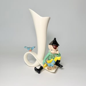 May include: A cream-colored ceramic vase with a whimsical design. It features a seated elf figure in green and yellow attire, a black hat, and boots. A blue bird rests on the vase's loop, and another sits near the elf. The vase has a tall, curved shape.