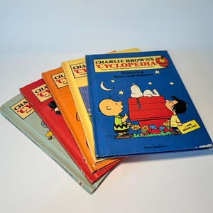 May include: A stack of colorful children's books titled "Charlie Brown's Cyclopedia." The books have covers in various colors, including blue, orange, red, and yellow. The blue book features Charlie Brown, Snoopy, and the text "Featuring Stars and Planets."