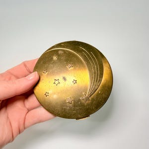 May include: A vintage, round, brass compact with a celestial design. The compact features a crescent moon and shooting star motif, along with several small stars. The brass has a warm, aged patina. The compact is held in a hand.