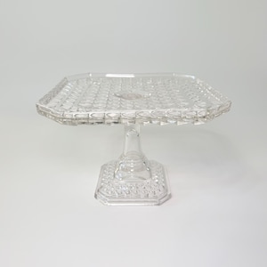 May include: A clear, square glass cake stand with a diamond pattern on the top and base. The stand has a pedestal base and a slightly raised edge. The design is vintage and elegant.
