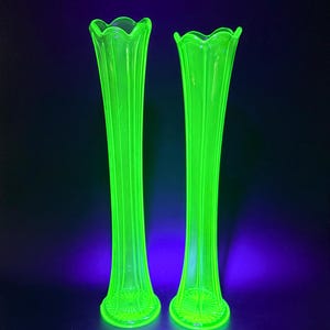 May include: A pair of tall, neon green glass vases. Each vase has a fluted design and a scalloped rim. The bases are detailed. The bright green colour contrasts with the dark background, highlighting their elegant, elongated form.