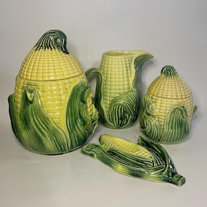 May include: A ceramic corn-themed kitchen set. The set includes a large lidded jar, a creamer pitcher, a small lidded jar, and a corn-shaped spoon rest. The pieces are yellow and green, with detailed corn husk and kernel designs.