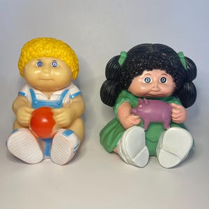 May include: Two Cabbage Patch Kids dolls. One doll has yellow hair, blue eyes, and is wearing a white and blue outfit and holding a red ball. The other doll has black hair, blue eyes, a green dress, and is holding a pink pig.