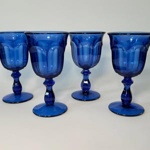 May include: Four cobalt blue wine glasses. Each glass has a rounded bowl with a scalloped rim, a detailed stem, and a circular base. The glasses are evenly spaced and arranged on a white surface.