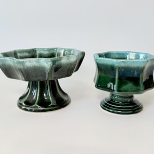 May include: Two ceramic pedestal bowls in shades of green. The bowls have a scalloped rim and sit on a rounded base. The larger bowl is taller than the smaller bowl. The bowls have a glossy finish.