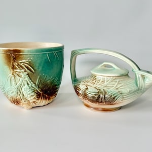 May include: A ceramic teapot and planter set. The teapot has a curved handle and a lid. The planter is a round bowl. Both pieces are teal, brown, and cream-colored with a textured, embossed design of leaves and branches.