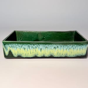 May include: A rectangular ceramic tray with a deep forest green interior and rim. The exterior features a reactive glaze effect, transitioning from a dark green top to a light blue middle and a pale yellow base with a textured, dripped appearance.