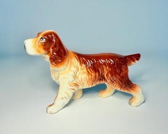 Peut inclure: Figurine de chien en céramique en position de marche. Le chien a une finition brillante avec une palette de couleurs blanche, marron et beige. La figurine est un objet décoratif, probablement pour l'exposition.