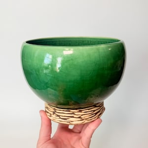 May include: A glossy, green ceramic bowl with a textured, tan base. The bowl has a rounded shape and a smooth, reflective surface. The base features a woven-like design, adding a contrasting texture to the piece. The bowl is held by a hand.