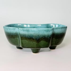 May include: A ceramic planter with a unique shape and a gradient color scheme. The planter transitions from a teal blue at the top to a deep green at the base. It rests on three small, white-tipped legs.