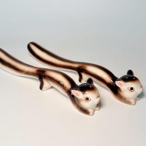 Peut inclure: Une paire de salières et poivrières en céramique en forme d'écureuils allongés. Chaque pièce présente un corps crème avec des rayures brun foncé le long du dos et de la queue, des oreilles teintées de rose et des trous sur le dessus de la tête.