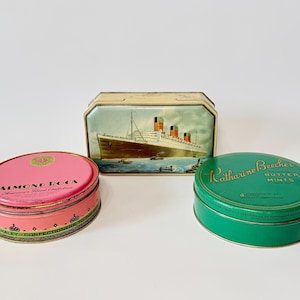 May include: Three vintage metal tins. A pink oval tin with "Almond Roca" text, a rectangular tin with a ship illustration, and a green round tin with "Katharine Beecher Butter Mints" text.